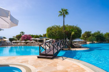 Kıbrıs 'ta ahşap köprüsü, berrak mavi suyu, palmiye ağaçları, pembe çiçekler, beyaz şemsiyeler, güneş panzeleri ve açık mavi bir gökyüzü olan bir tatil yeri..