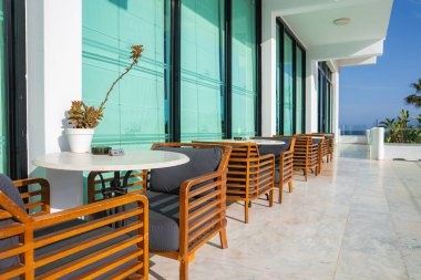 Koyu yastıklı modern ahşap sandalyeler ve mermer terastaki yuvarlak beyaz masalar, cam pencereler, palmiye ağaçları ve deniz manzarası ile çevrili..