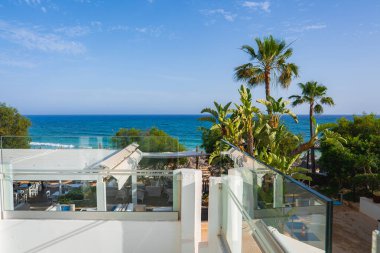 Palmiye ağaçları, tropikal bitkiler ve arka planda Akdeniz 'in bulunduğu açık hava oturma alanlarına bakan modern bir teras..