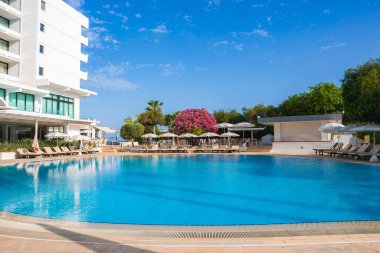 Kıbrıs 'ta el değmemiş bir havuzu, güneş pansiyonları, şemsiyeleri, pembe begonvilleri, yemyeşil bir oteli ve arka planda açık mavi bir gökyüzü olan modern bir otel..