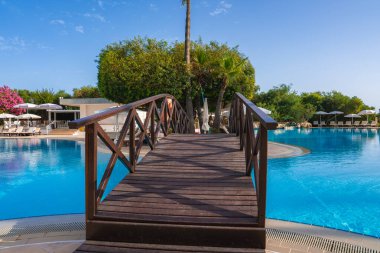 Kıbrıs 'ta, palmiye ağaçları, çiçek açan bitkiler, güneş pansiyonları ve açık mavi gökyüzü ile çevrili büyük bir havuz üzerinde ahşap bir köprü bulunan sakin bir tatil köyü..