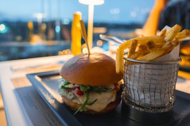 Eritilmiş peynirli, yeşillikli ve domatesli gurme bir burger. Modern bir restoranda, yumuşak akşam ışıklarıyla birlikte siyah bir tepside servis ediliyor..