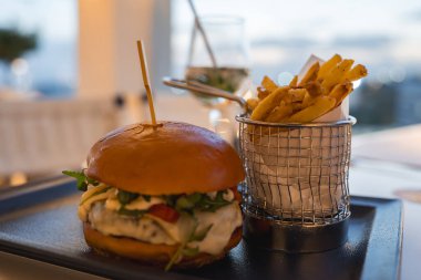Sebzeli, peynirli ve domatesli gurme burger ve patates kızartması. Limonlu su bardağı yumuşak ışıklı, lüks bir yemek alanında durur..