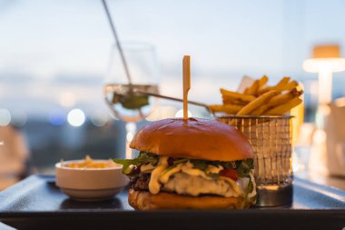 Sebzeli, peynirli ve siyah tepside patates kızartması ve soslu gurme burger. Sıcak bir akşam atmosferinde bir bardak beyaz şarap oturur..