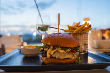 Taze yeşillik, domates ve patates kızartması ve sos ile servis edilen kremalı bir gurme burger. Bir kadeh şarap dingin akşam arkaplanını tamamlıyor..