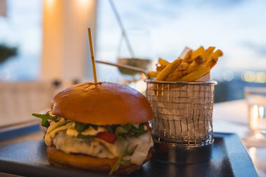 Meyveli, yeşil, peynirli ve domatesli gurme burger. Metal bir sepetin içinde patates kızartmasıyla servis ediliyor. Yumuşak ışıklandırma ve bulanık açık hava görünümü.