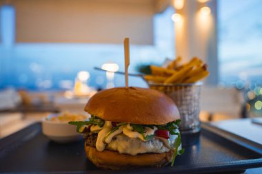 Eritilmiş peynir, yeşillik ve siyah tepside soslu kızarmış patates ve lahana salatasıyla servis edilen gurme burger. Alacakaranlık şehir manzarası arka planda görünür.