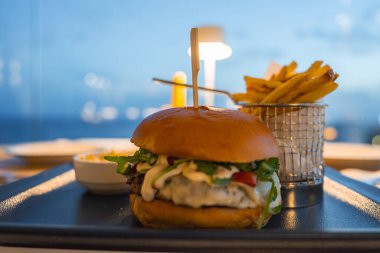 Taze sebzeli, domatesli ve siyah tepside kremalı soslu gurme burger ve patates kızartması. Arka planda bulanık deniz ve akşam gökyüzü.