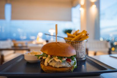 Sebzeli, peynirli ve soslu bir gurme burger akşam atmosferi olan bir açık hava restoranında siyah bir tepside patates kızartması ve lahana salatası ile servis edilir..
