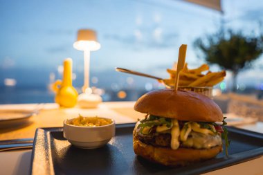 Sebzeli gurme burger ve siyah tabakta sos. Patates kızartması ve sosla servis ediliyor. Deniz manzaralı ve sıcak akşam aydınlatmalı açık hava yemek alanı..