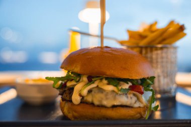 Eritilmiş peynirli, yeşillikli ve soslu bir gurme burgerin kapağı açık hava yemek alanında kızarmış patates ve sosla servis ediliyor..