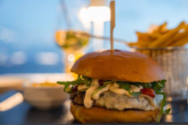 Erimiş peynir, yeşillik ve çöreğin üzerine sos konmuş gurme bir burger yakın çekim. Arkadaki patates kızartması ve beyaz şarap güzel bir yemek olduğunu gösteriyor..