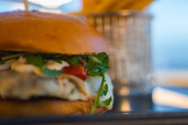 Roka, peynir ve domatesli altın hamburgerle metal bir sepetin içinde patates kızartması. Kıbrıs 'ta hafif ışıklandırma ve kıyı manzarası.