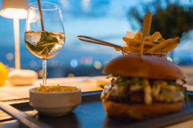 Taze malzemeli bir hamburger, metal sepette patates kızartması, lahana salatası ve akşam bulanık bir şehir manzarasına karşı naneli içecek..