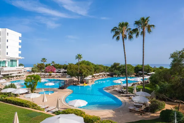 Kıbrıs Rum Kesimi 'nde bir havuz, güneş şezlongları, şemsiyeler, yüksek palmiye ağaçları, yemyeşil yemyeşil, modern beyaz bir otel ve arka planda deniz yer alıyor..
