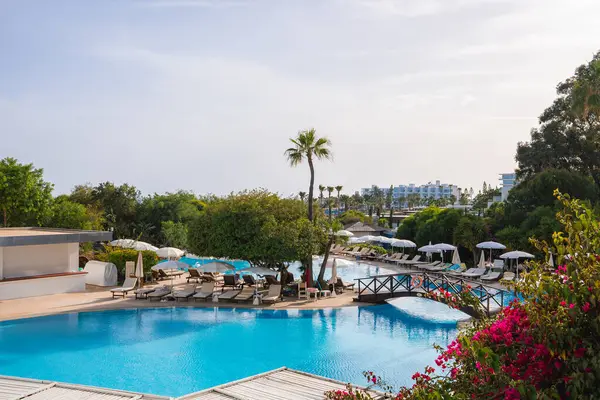 Kıbrıs 'ta bir tatil beldesinde büyük bir havuz, ahşap bir köprü, güneş pansiyonları, şemsiyeler, yemyeşil çiçekler ve modern otel binaları yer alıyor..