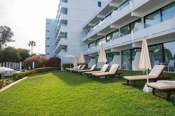 Birçok balkonlu, güneşli bahçeli, güneşli, şemsiyeli, çiçekli çalıları ve uzun bir palmiye ağacıyla çevrili beyaz, lüks bir otel..