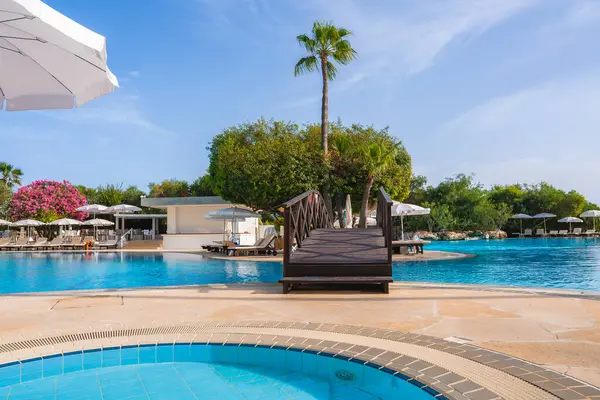 Kıbrıs 'ta bir tatil köyünde ahşap bir köprü, beyaz şemsiyeli güneş panzeleri, yemyeşil, pembe çiçekli ağaçlar ve uzun bir palmiye ağacı yer alıyor..