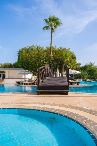 Kıbrıs 'ta bir tatil köyünde ahşap bir köprü, uzun bir palmiye ağacı, beyaz şemsiyeler, güneşli havuz ve güneşli gökyüzünün altında daha küçük dairesel bir havuz yer alıyor..