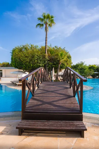 Bir tahta köprü, palmiye ağaçları, tropik bitkiler, beyaz binalar ve şemsiyelerin altındaki şezlonglarla çevrili bir Kıbrıs tatil beldesindeki berrak mavi havuzu geçer..