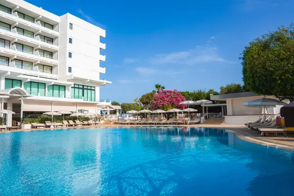 Kıbrıs 'ta modern beyaz bir otel, güneş pansiyonları, şemsiyeler, pembe bougainvillea, yemyeşil ve açık mavi gökyüzü olan el değmemiş bir havuza bakıyor..