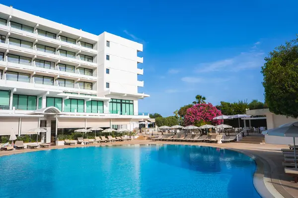 Kıbrıs 'ta balkonları, büyük bir havuzu, güneşlik, şemsiyeleri, pembe çiçekli bir ağacı ve açık mavi bir gökyüzü olan modern beyaz bir otel..