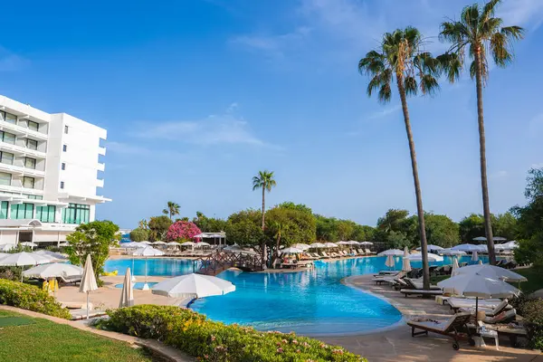 Kıbrıs 'ta geniş bir havuzu, güneşli odaları, şemsiyeleri, uzun palmiye ağaçları, yemyeşil bir bahçesi ve arka planda açık mavi bir gökyüzü olan modern beyaz bir otel..