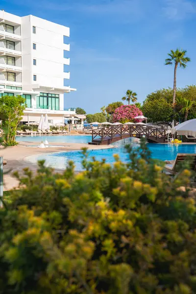 Kıbrıs 'ta bir tatil beldesinde ahşap köprüsü, yemyeşil yeşilliği, pembe çiçekli ağaçları ve berrak mavi gökyüzünün altında modern beyaz bir otel yer alıyor..