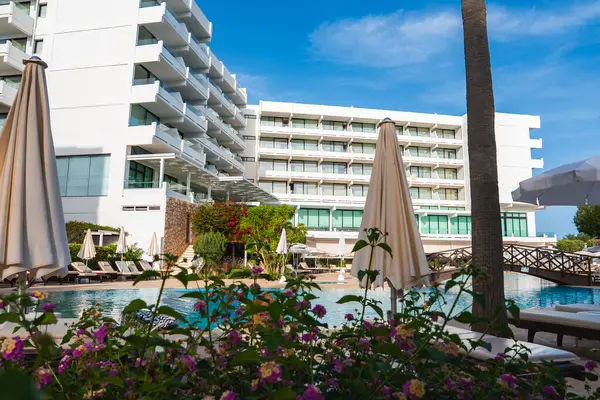 Balkonları, şezlongları, çiçekli bitkileri, ahşap bir köprüsü ve uzun bir palmiye ağacıyla çevrili bir havuzu olan modern beyaz çok katlı bir otel..