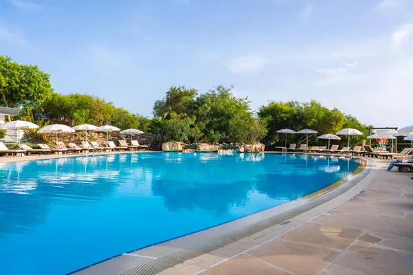 Berrak mavi sularla çevrili dingin bir yüzme havuzu, yemyeşil, güneşli şezlonglar, şemsiyeler ve Kıbrıs tatil beldesinde taş döşeli bir güverte..