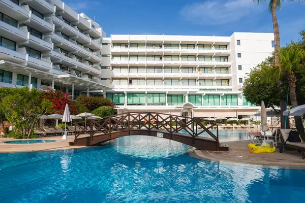 Balkonları olan modern beyaz bir otel ahşap köprüsü olan bir havuza bakıyor. Palmiye ağaçları, güneş pansiyonları, şemsiyeler ve canlı bitkilerle çevrili..