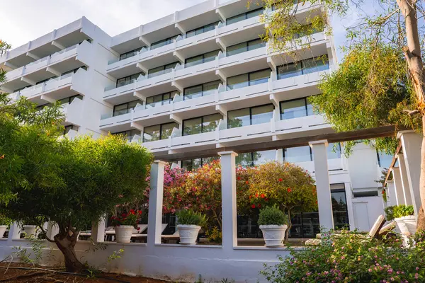 Camdan balkonları olan çok katlı beyaz bir otel, beyaz saksılarda çiçek açan bitkilerle dolu bir bahçe ve Akdeniz 'de geçen bir pergola yapısı..