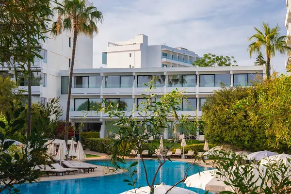 Kıbrıs 'taki modern beyaz otel binası yeşil ve palmiye ağaçları ile çevrili, dingin bir havuzu, güneş pansiyonları, şemsiyeleri ve açık mavi bir gökyüzü var..