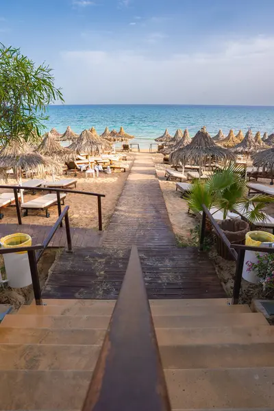 Ahşap bir patika, sazdan şemsiyeler ve güneş şezlonglarıyla kaplı kumlu bir sahile çıkıyor. Turkuaz deniz ve yemyeşil yemyeşil tropik bir ortam yaratır..