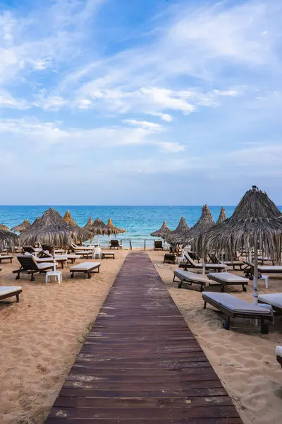 Kıbrıs 'taki sakin bir plajda ahşap bir yürüyüş yolu, turkuaz deniz, altın kum, güneş panjurları, saman şemsiyeler ve açık mavi bir gökyüzü yer alıyor..