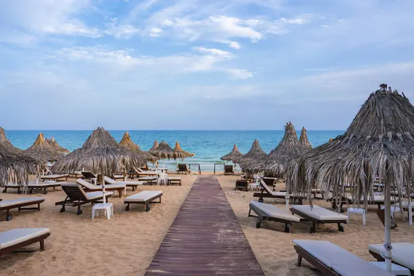 Kıbrıs 'taki kumlu bir plajda, parçalı bulutlu bir gökyüzünün altında ahşap bir yürüyüş yolu, sıra sıra güneş şezlongları, sazdan şemsiyeler ve sakin turkuaz bir su bulunuyor..