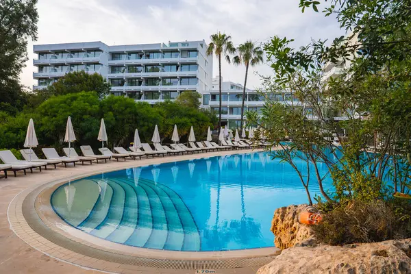 Yarı yuvarlak basamaklı kavisli bir yüzme havuzu, beyaz şemsiyeler ve palmiye ağaçları ve Kıbrıs 'ta modern beyaz bir otel çerçeveli şezlonglar..