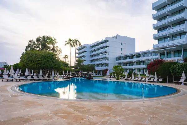 Büyük yuvarlak bir havuz, şezlonglar, şemsiyeler ve dekoratif bir köprü, palmiye ağaçları ile çevrili ve modern beyaz otel binaları tarafından gözden kaçırılmış..