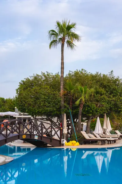 Kıbrıs 'taki lüks bir tatil köyünde ahşap bir köprü, sarı şişme ördek, palmiye ağaçları, beyaz şemsiyeler ve huzurlu bir ortamda şezlonglar yer alıyor..