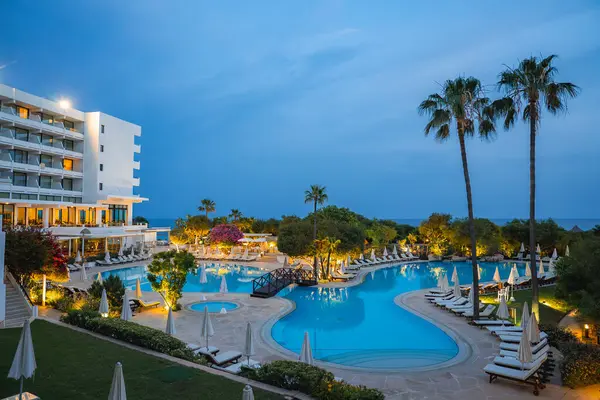 Kıbrıs 'ta ışıklı bir yüzme havuzu, şezlonglar, palmiye ağaçları, bougainvillea, modern bir otel ve arka planda deniz bulunan bir tatil beldesinin akşam manzarası.