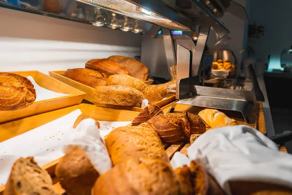 Sıcak aydınlatma altında ahşap tepsilerde çeşitli somunlar, baget ve rulo çeşitleri, modern bir yemek alanında, paslanmaz çelik ekipman ile ayarlanmış.