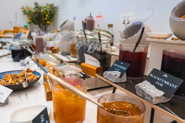 Üzerinde kırmızı çilek kavanozları, kuşüzümü reçelleri, bal, granola ve beyaz bir tezgahta baharatlar bulunan canlı bir kahvaltı sergisi..