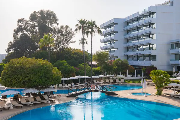 Balkonları, büyük bir havuzu, güneş pansiyonları, şemsiyeleri, ahşap bir köprüsü, yemyeşil bir bahçesi ve Kıbrıs 'taki yüksek palmiye ağaçları olan modern beyaz tatil köyü binası..