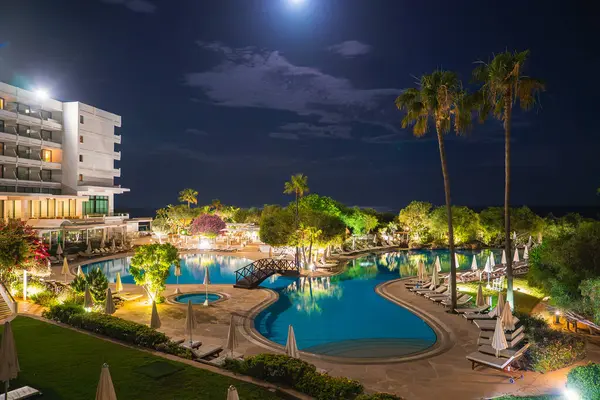 Kıbrıs 'ta mehtaplı gökyüzünün altındaki bir tatil köyünde şezlonglar, şemsiyeler ve palmiyelerle çevrili, ahşap bir köprüyle aydınlatılmış büyük bir yüzme havuzu..