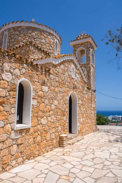 Kemerli camları ve muhtemelen St. Elias Kilisesi 'ne benzeyen bir çan kulesi olan taştan bir kilise, Kıbrıs' ta açık mavi gökyüzü altında bir kıyı şeridine ve binalara kurulmuş..