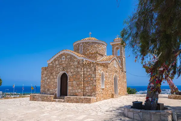 Kıbrıs 'taki Aziz Elias Kilisesi' nde Bizans mimarisi, süslenmiş bir kurdele ağacı ve parlak mavi gökyüzü altında Akdeniz manzarası yer alıyor..
