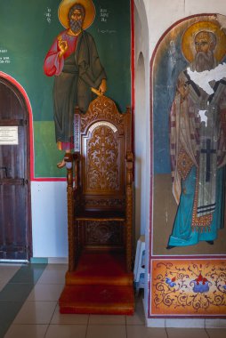 Karmaşık oyulmuş ahşap bir sandalye, azizlerin canlı freskleri, koyu ahşap bir kapı ve St. Elias Kilisesi 'nin iç döşemelerinin cilalı olduğu bir duvara karşı oturur..