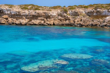 Görünür sualtı kayalarıyla kristal berrak turkuaz sular, Kıbrıs 'ın kıyı şeridi boyunca uzanan seyrek bitki örtüsüyle kaplı engebeli açık renkli uçurumlarla buluşuyor.