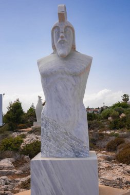 Başlık gibi bir miğferi olan biçimlendirilmiş bir insan figürünün mermer heykeli Kıbrıs 'ta kayalık, benekli bir arazide taş bir kaide üzerinde duruyor..