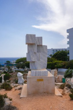 Modern geometrik mermer heykel, plaketli bir kaide üzerinde, açık deniz kenarına kurulmuş. Arka planda lüks bir otel ve kayalık bir arazi görünüyor.